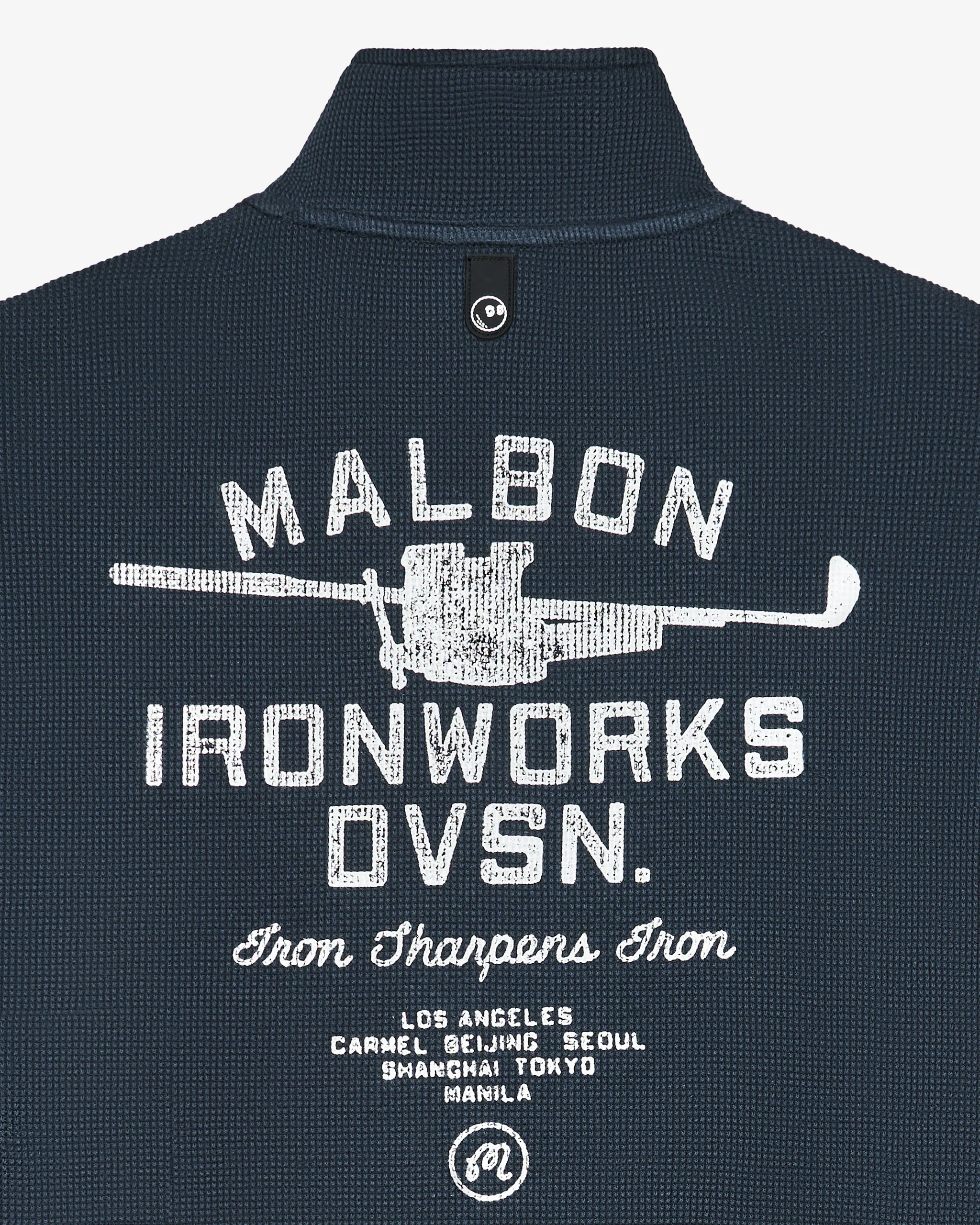Close-up of the back of a dark navy textured polo shirt with a white distressed graphic reading 'MALBON IRONWORKS DVSN.' and additional text below, on a light gray background.