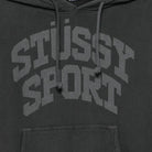 Close-up of a dark charcoal gray hoodie featuring a large, tonal gray 'STUSSY SPORT' graphic printed across the chest, with matching drawstrings and a front kangaroo pocket.