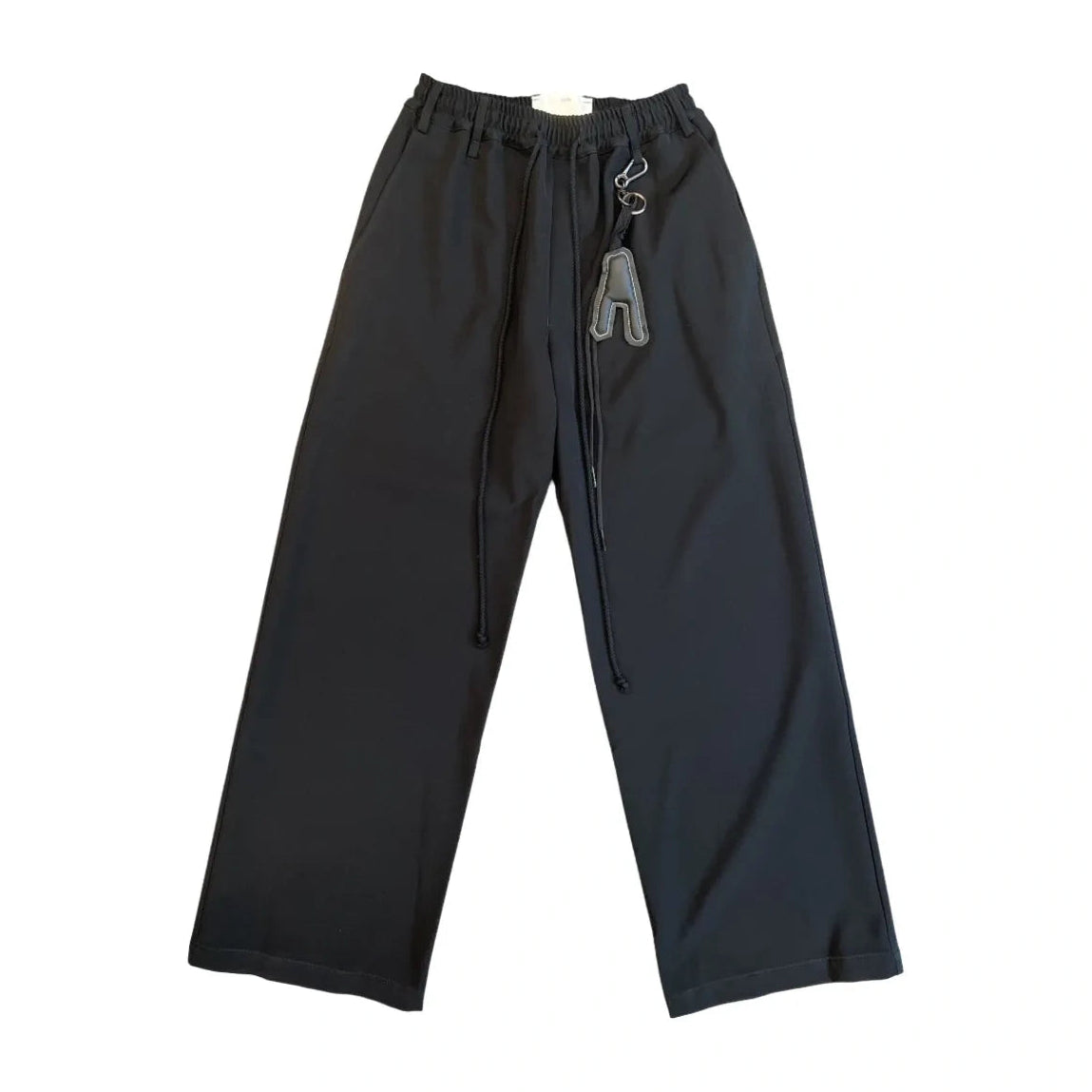 Front view of black wide-leg Song For The Mute pants laid flat. They have an elastic drawstring waist with long rope cords and a carabiner key charm with an “A” tag hanging from a belt loop.