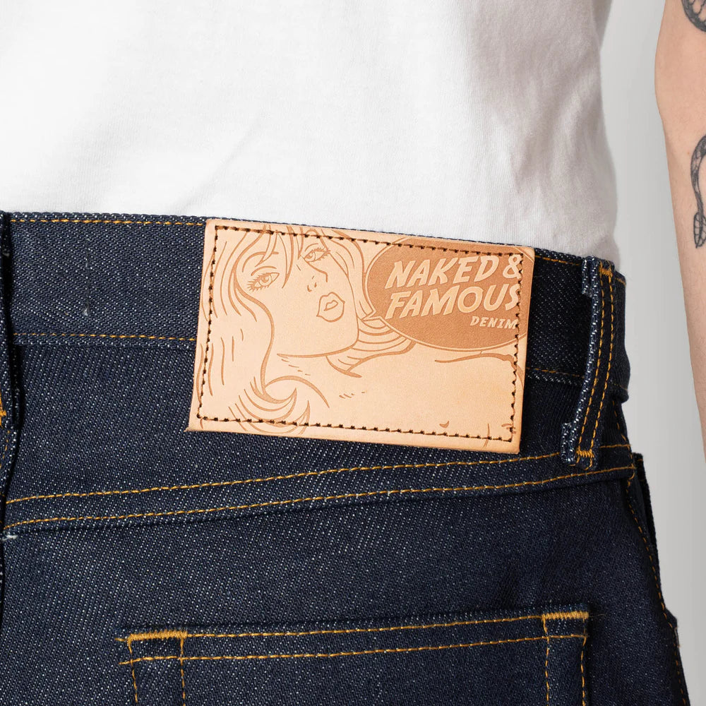 Naked & Famous denim label on a pair of jeans with a white shirt and tattooed arm in the background.