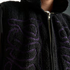 Black fleece jacket with purple embroidery on a neutral background