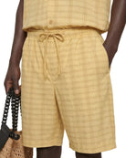 Person wearing a yellow plaid outfit with shorts and a top, holding a woven bag.
