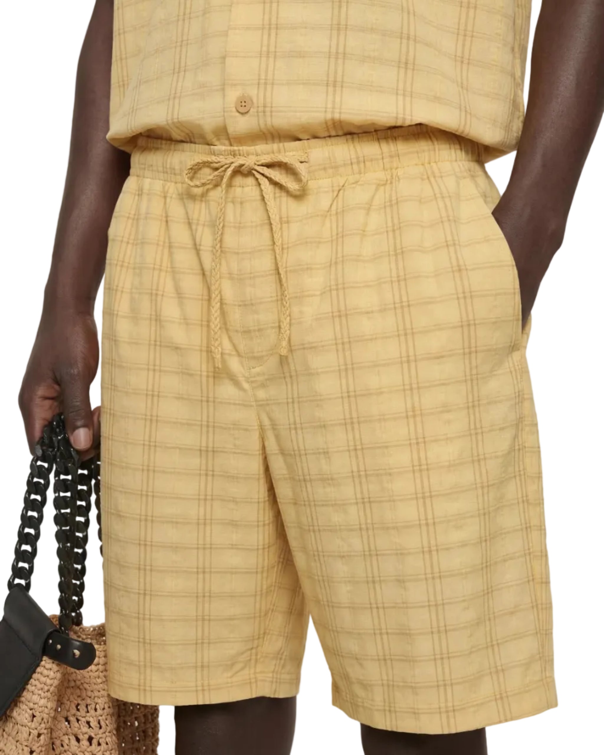 Person wearing a yellow plaid outfit with shorts and a top, holding a woven bag.