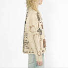 A side-profile view of a model with long blonde curly hair wearing a cream-colored graphic jacket with black text and illustrations, paired with light blue jeans, against a plain white background.