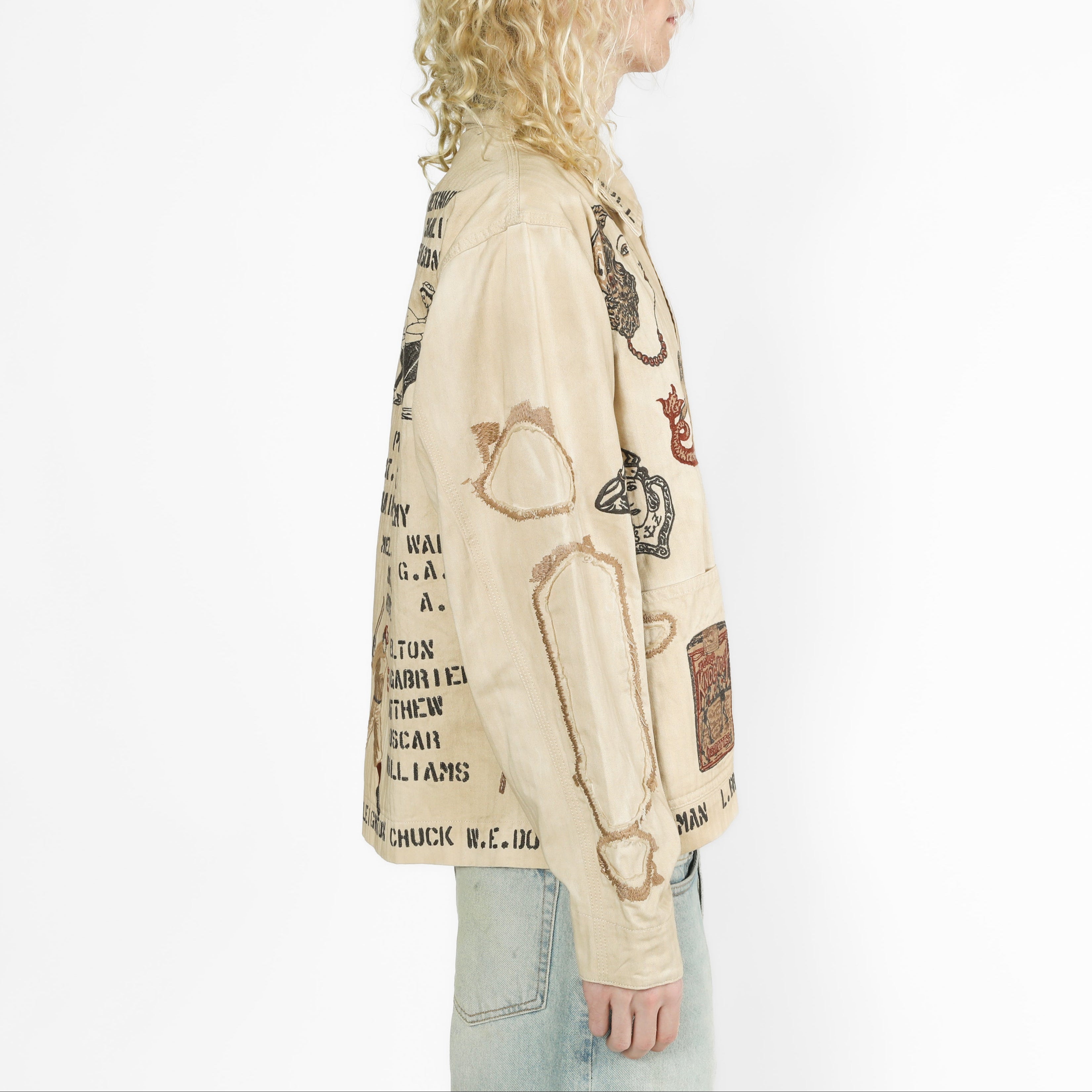 A side-profile view of a model with long blonde curly hair wearing a cream-colored graphic jacket with black text and illustrations, paired with light blue jeans, against a plain white background.