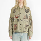 A model with long blonde hair wearing a faded olive chore jacket with multiple embroidered images all over.