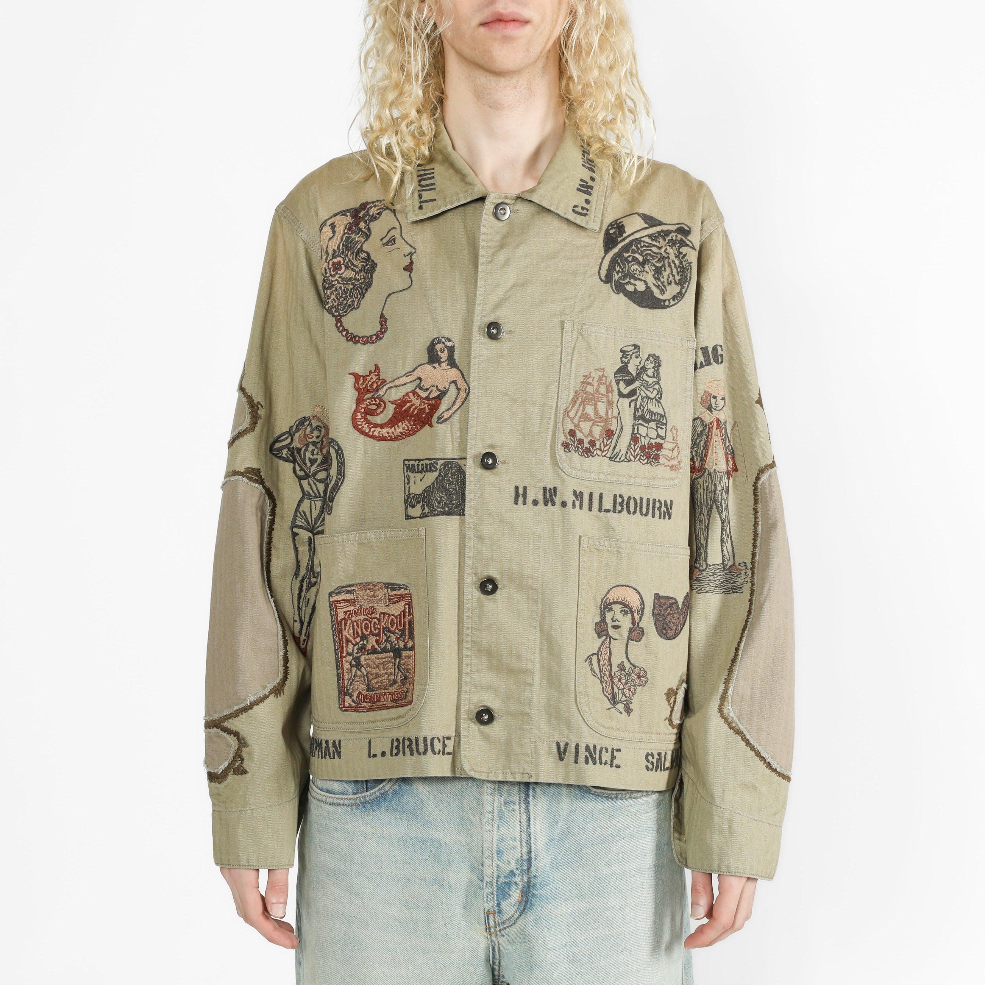 A model with long blonde hair wearing a faded olive chore jacket with multiple embroidered images all over.