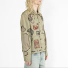 A model with curly blonde hair is shown from the side, wearing a beige button-up jacket with a vintage collage print of illustrated faces, postcards, and text, paired with light blue jeans against a plain white background.