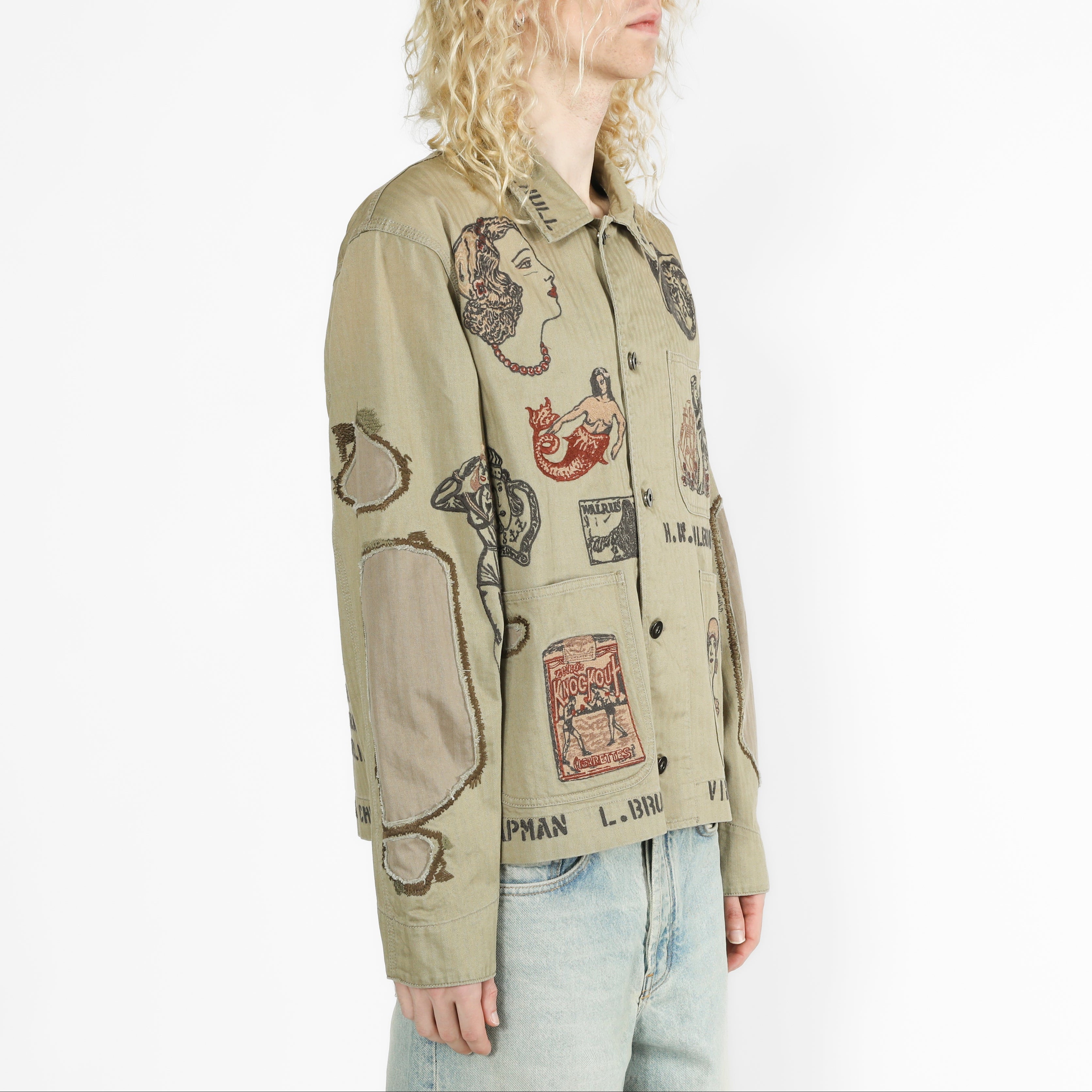 A model with curly blonde hair is shown from the side, wearing a beige button-up jacket with a vintage collage print of illustrated faces, postcards, and text, paired with light blue jeans against a plain white background.