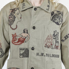 Close-up of a person wearing a light olive-beige button-front shirt jacket with large embroidered vintage-style illustrations and text, including a mermaid, a woman in profile, a ship, and the words H.W.