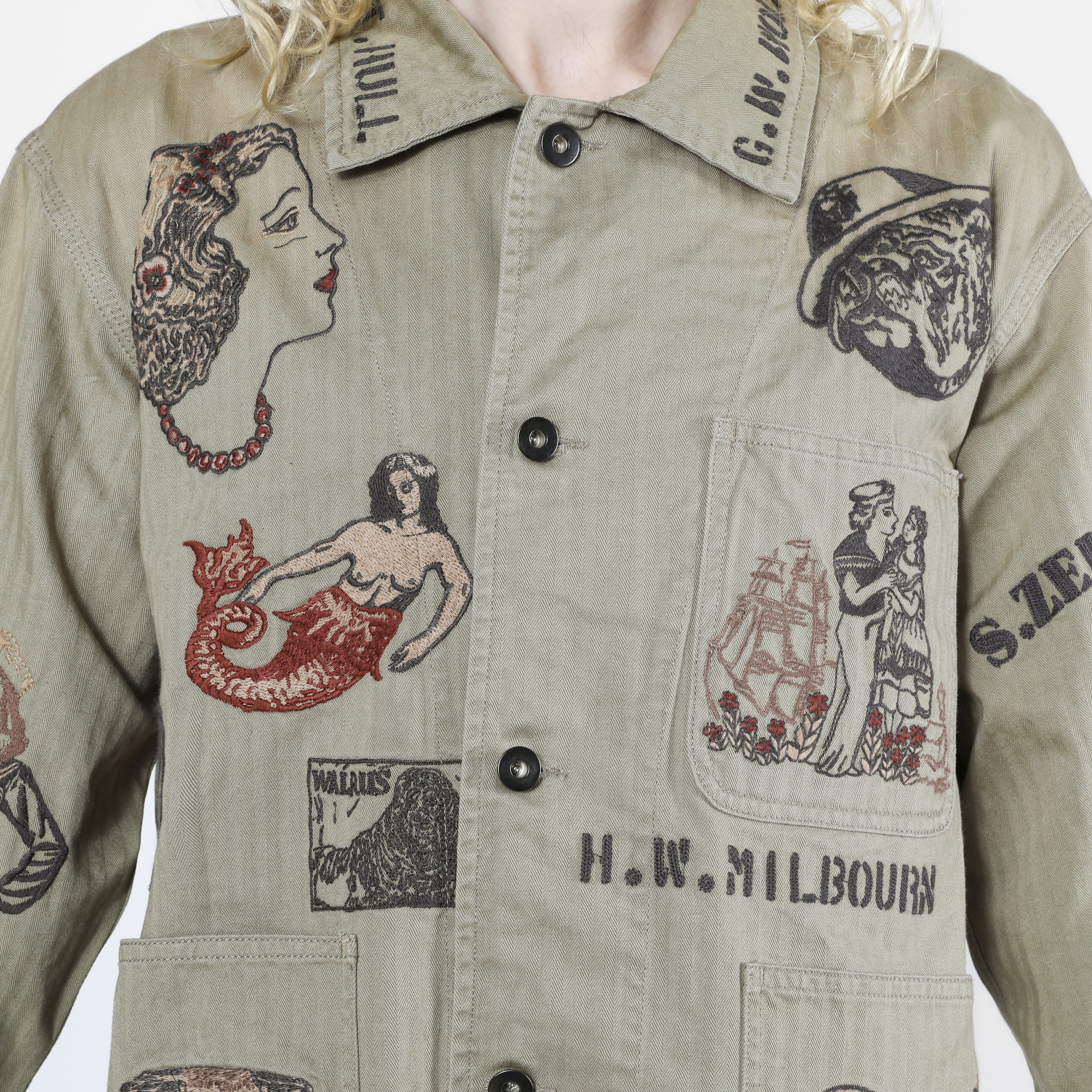 Close-up of a person wearing a light olive-beige button-front shirt jacket with large embroidered vintage-style illustrations and text, including a mermaid, a woman in profile, a ship, and the words H.W.