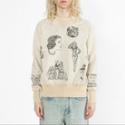 A model with long blonde hair wearing a beige crewneck sweatshirt with black vintage-style graphic prints and light blue jeans, set against a plain white studio background.