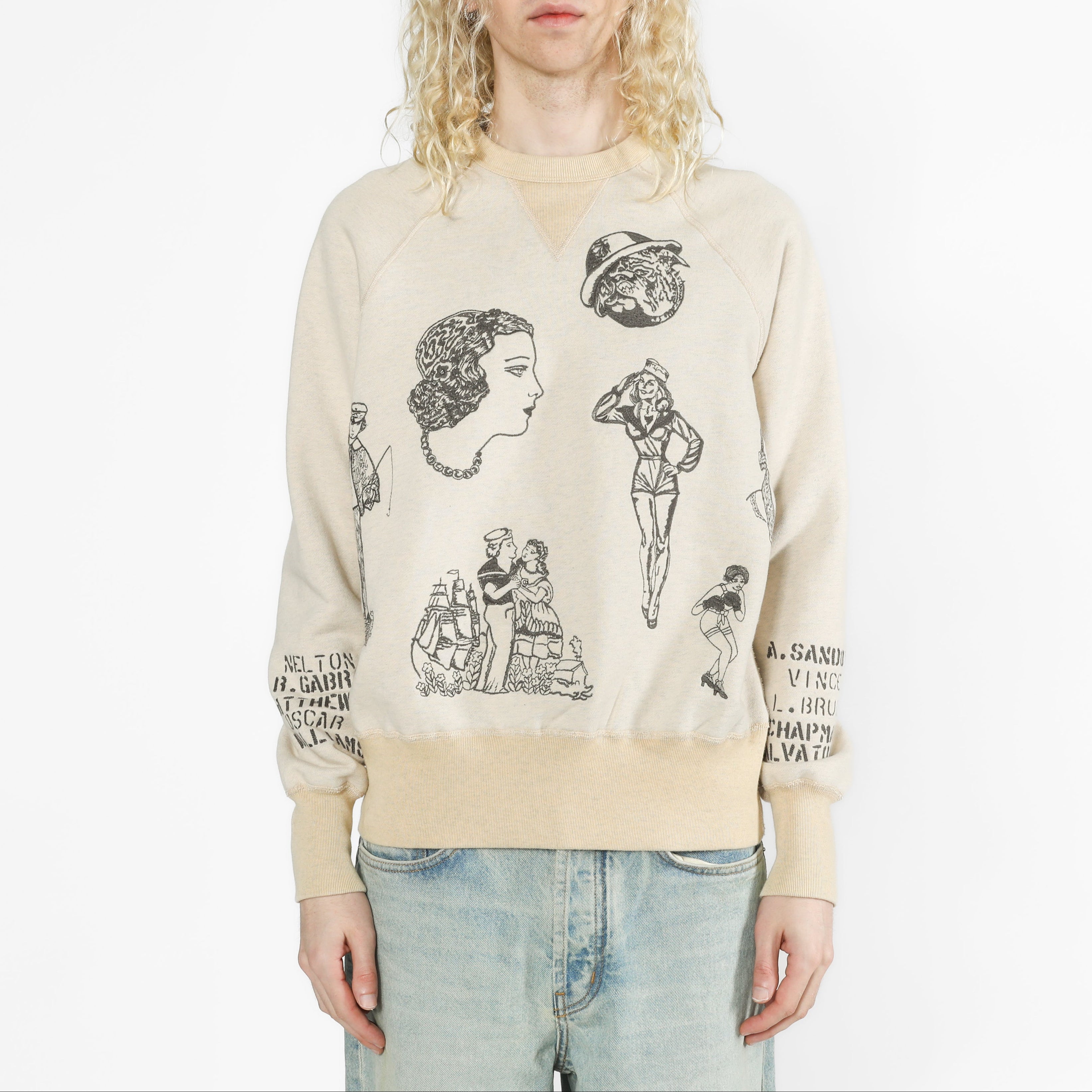 A model with long blonde hair wearing a beige crewneck sweatshirt with black vintage-style graphic prints and light blue jeans, set against a plain white studio background.