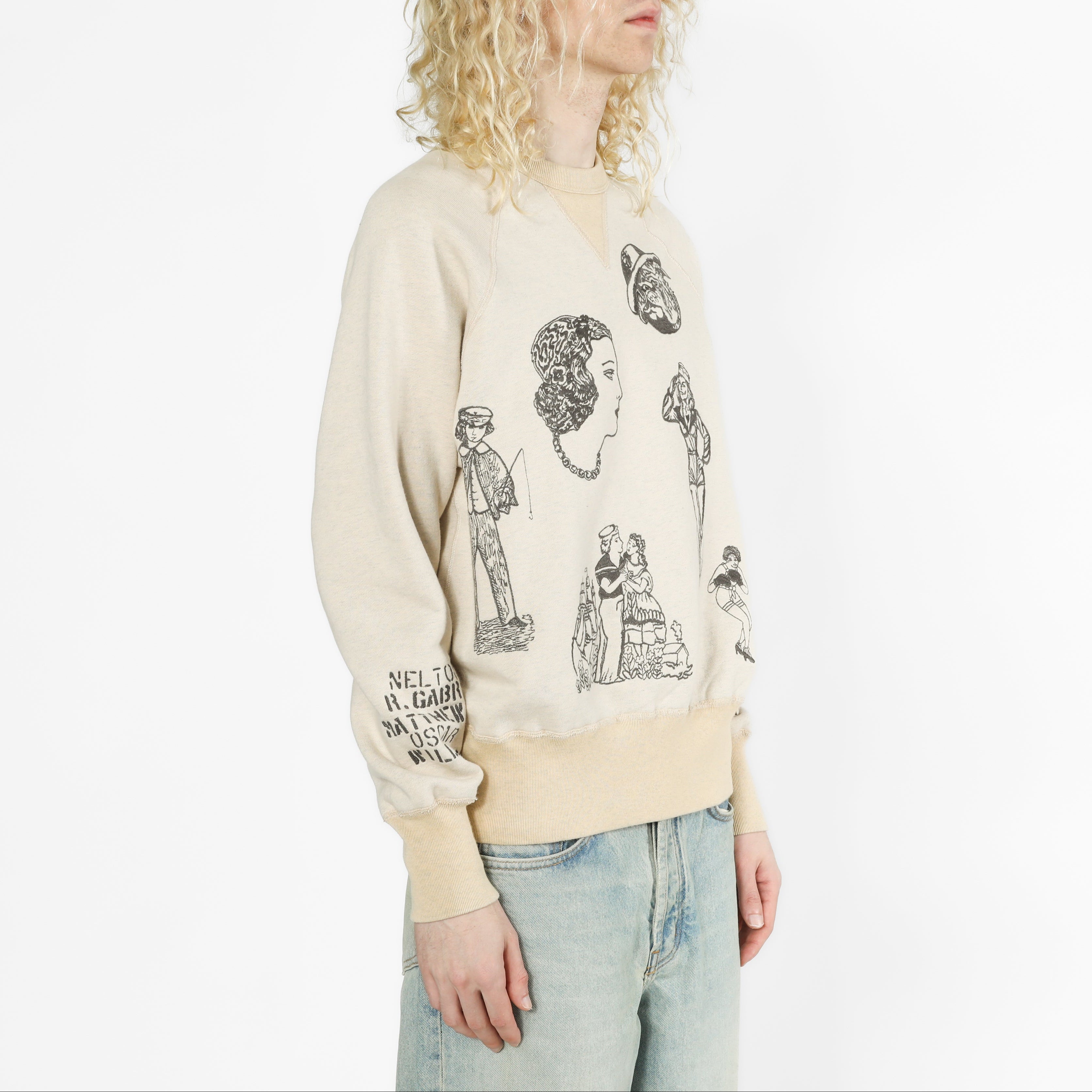 A model with curly blonde hair wearing a cream-colored crewneck sweatshirt with black graphic illustrations of historical figures and light-wash blue jeans, photographed against a plain white studio background.