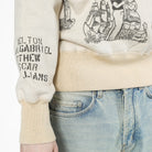 Close-up of a cream-colored crewneck sweatshirt with black graphic prints on the sleeve and chest, paired with light-wash blue jeans.