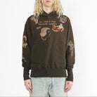 A model with long blonde hair wearing a dark brown hoodie with gothic graphic prints and text, paired with light-wash jeans, against a plain white studio background.