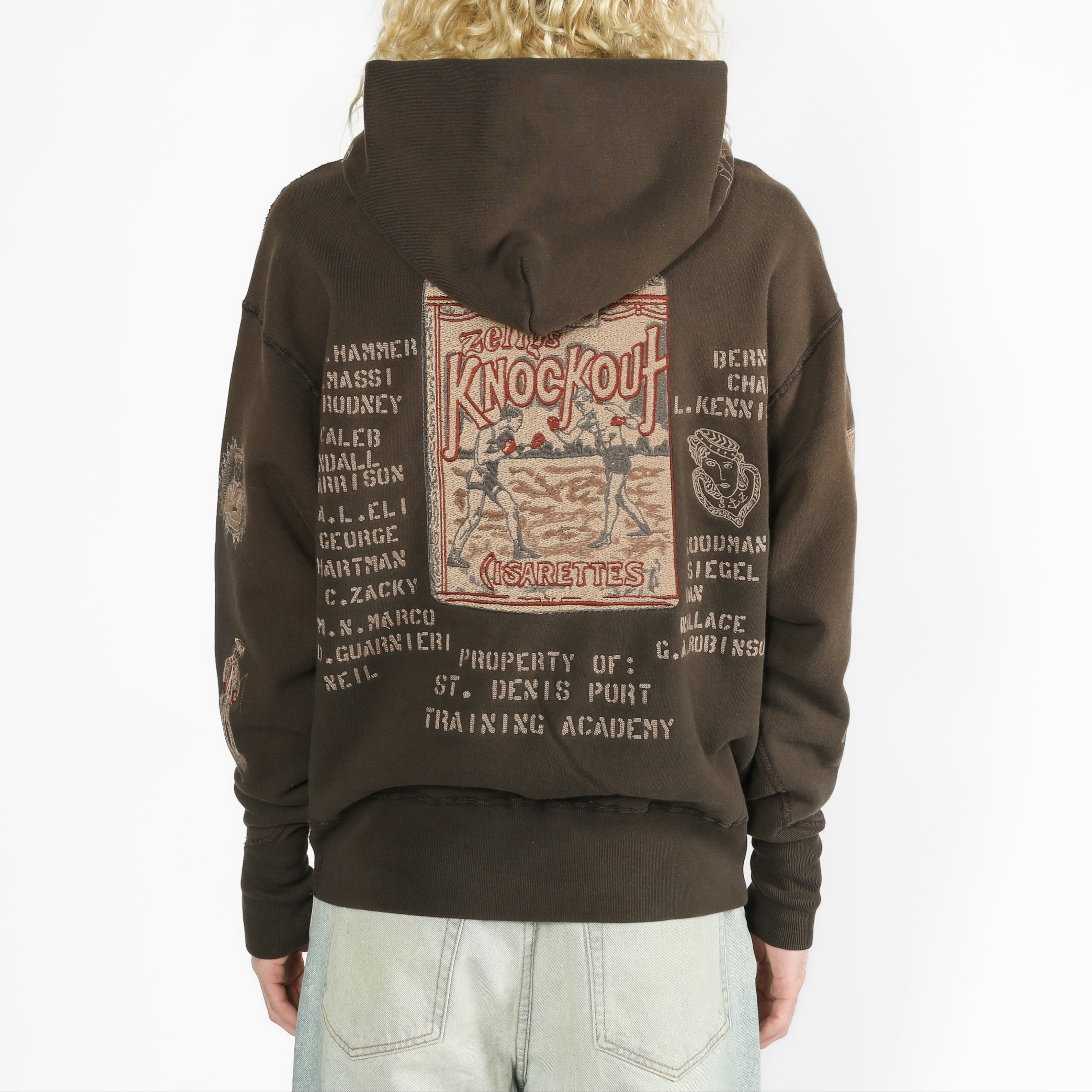 A person shown from the back wearing a dark brown hoodie with a large vintage-style graphic print on the back. The hoodie has distressed detailing and text elements in muted red, beige, and white.