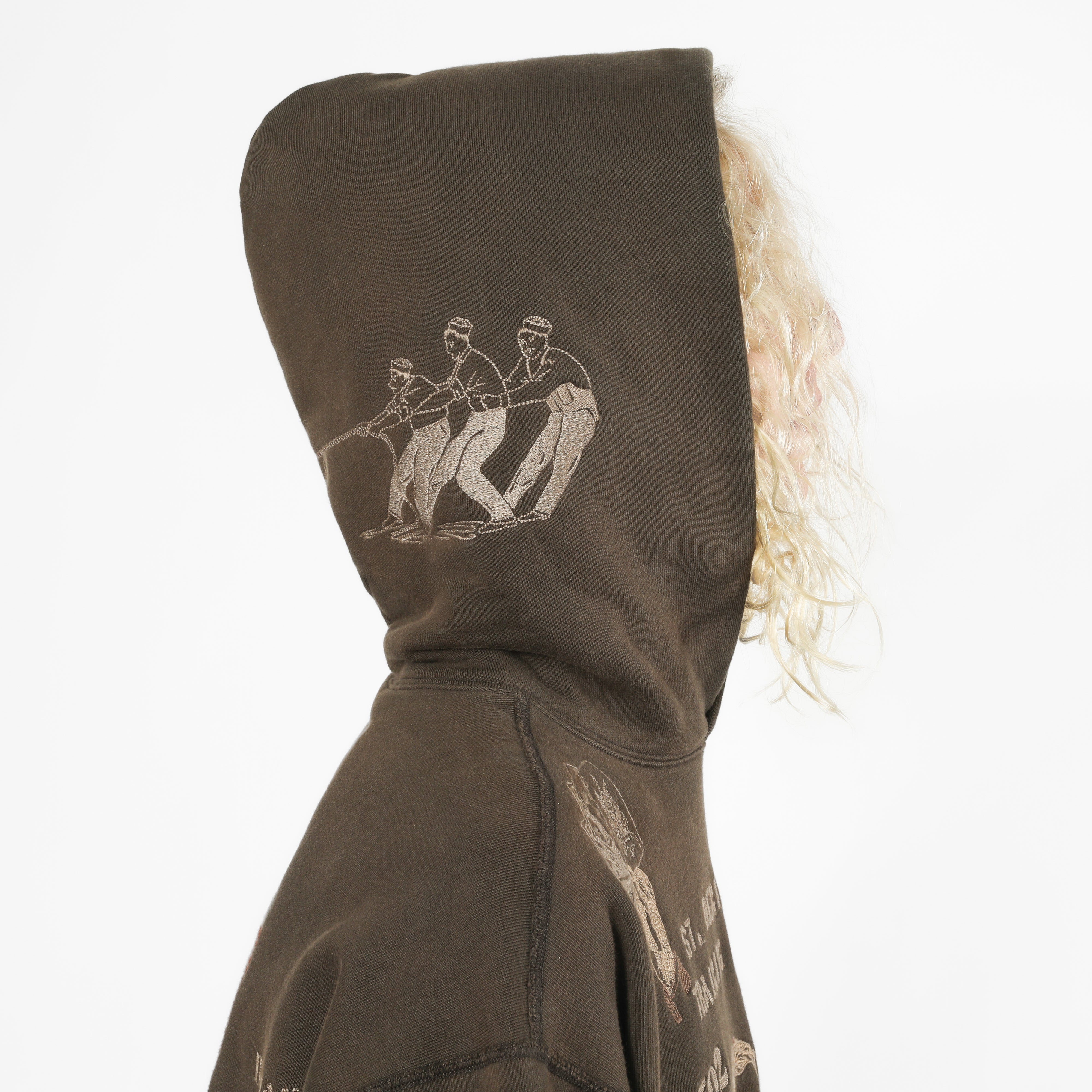A close-up side view of a person wearing a dark brown hooded sweatshirt with a light beige graphic print of three figures on the hood, against a plain white background.