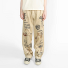 A model stands against a plain white studio background wearing a cream-colored oversized short-sleeve t-shirt and beige wide-leg pants with embroidered vintage-style print, paired with black and white sneakers.