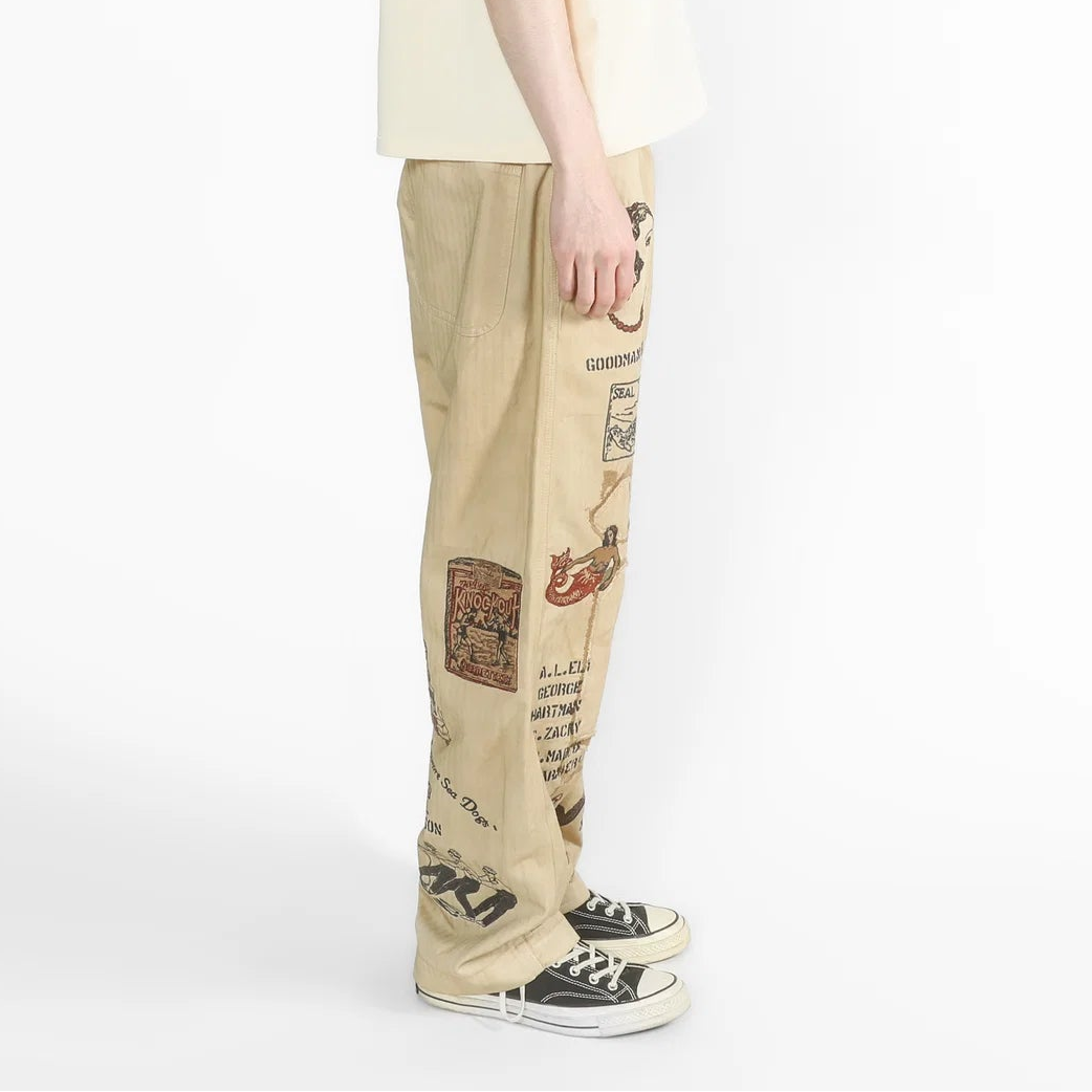 A model shown from the side profile wearing a cream-colored oversized t-shirt, beige graphic pants, and black canvas sneakers against a plain white studio background.