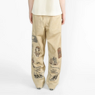 A model viewed from the back wearing a cream-colored short-sleeve t-shirt and beige wide-leg pants with graphic prints on the legs, set against a plain white studio background.