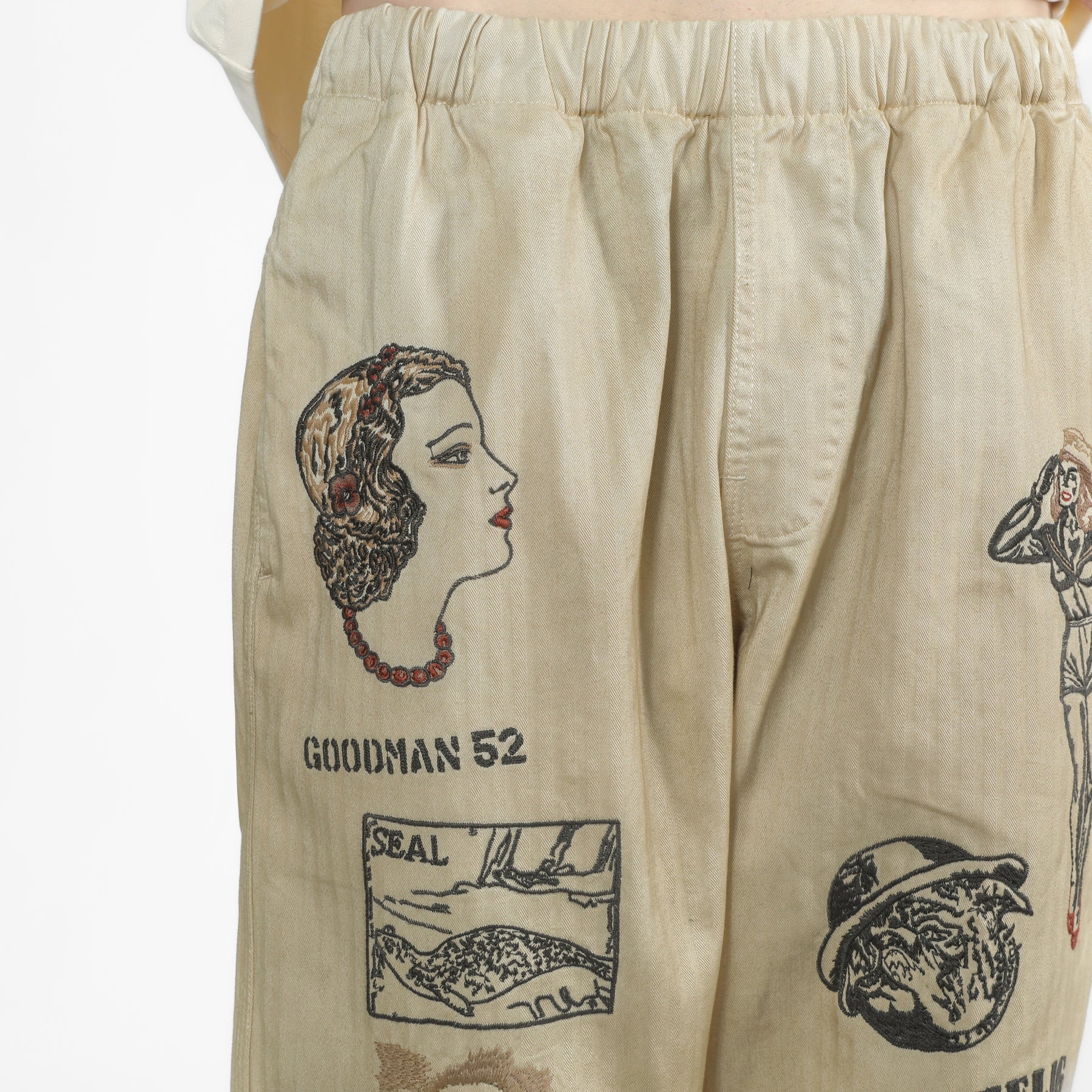 Beige herringbone pants featuring vintage-style embroidered illustrations and text including 'GOODMAN 52' and 'SEAL'.