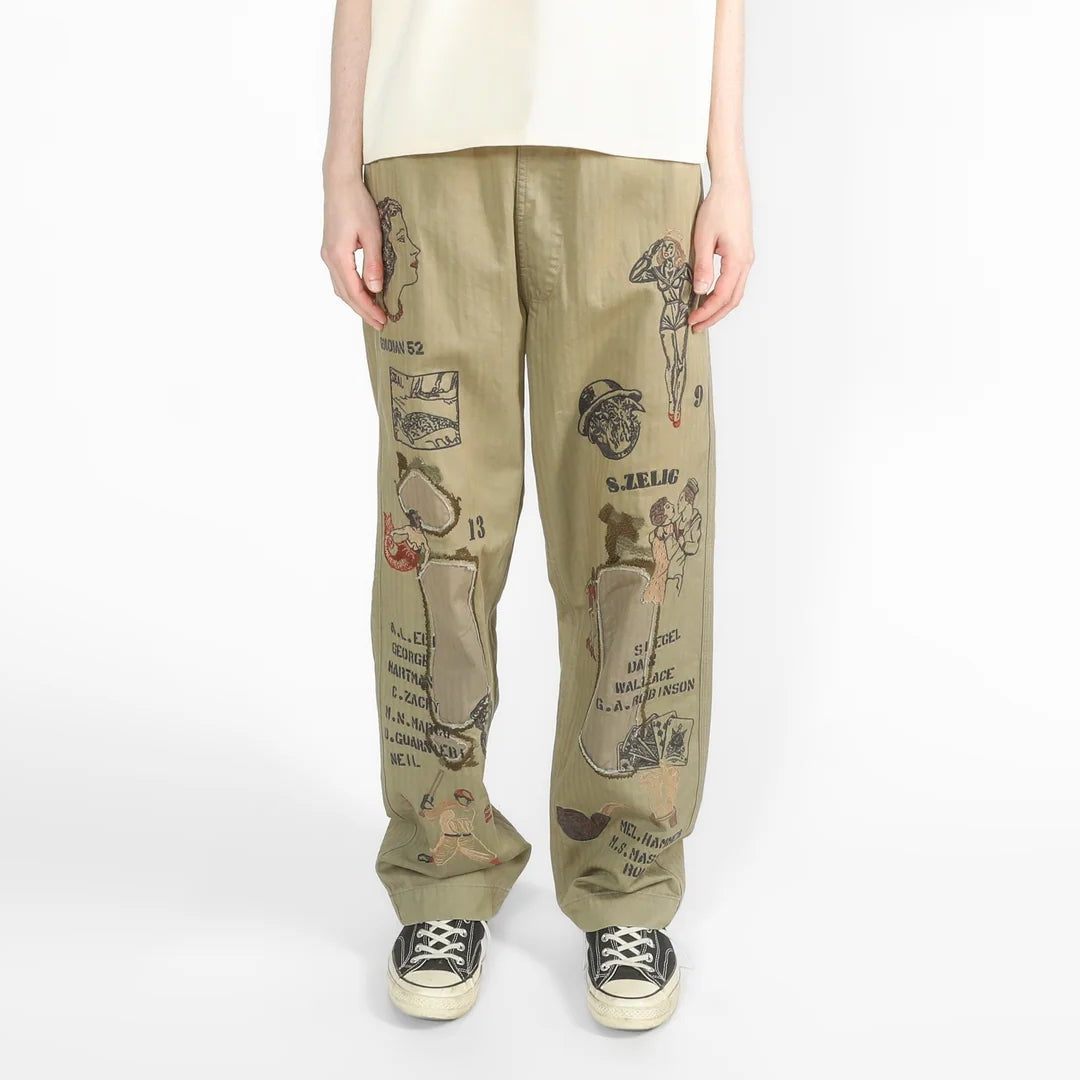 A model stands against a plain white studio background wearing a cream-colored oversized short-sleeve t-shirt, olive green wide-leg pants with black illustrated graphics, and black-and-white sneakers.