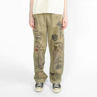 A model stands against a plain white studio background wearing a cream-colored oversized short-sleeve t-shirt, olive green wide-leg pants with black illustrated graphics, and black-and-white sneakers.