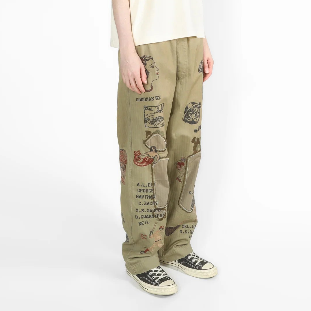 A model stands in profile wearing a cream-colored oversized t-shirt, olive green graphic cargo pants, and black-and-white sneakers against a plain white studio background.