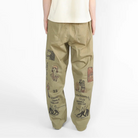 A model viewed from behind wearing a cream-colored short-sleeve t-shirt and olive green cargo pants with black graphic prints, set against a plain white studio background.