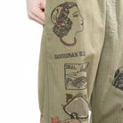 Close-up of a pair of olive green pants with vintage-style embroidered graphics and text including 'GOODMAN 52' and 'SEAL'.