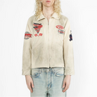 A model with long blonde hair wearing a beige zip-up jacket with multiple red, black, and white patches, paired with light blue jeans, against a plain white background.