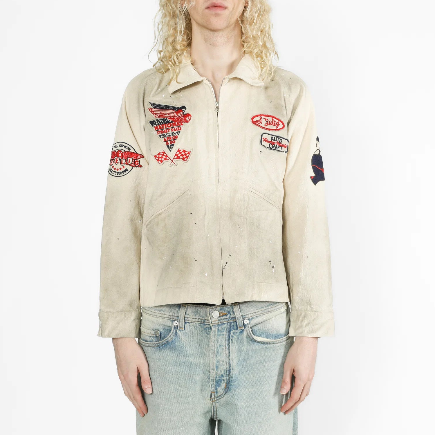 A model with long blonde hair wearing a beige zip-up jacket with multiple red, black, and white patches, paired with light blue jeans, against a plain white background.