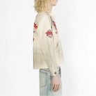 A model with long blonde curly hair is shown in profile wearing a heavily distressed off-white jacket with red and black graphic patches on the sleeves, paired with light-wash blue jeans against a plain white background.