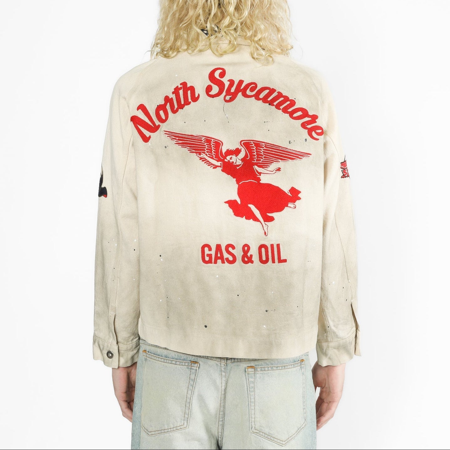 A model viewed from behind wearing a heavily distressed cream-colored jacket with a large red graphic on the back reading 'North Sycamore GAS & OIL' and featuring a winged figure. The model is also wearing light-wash jeans against a plain white background.