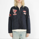 A model with long blonde hair wearing a dark navy blue utility jacket with multiple embroidered patches, paired with light-wash jeans, against a plain white studio background.