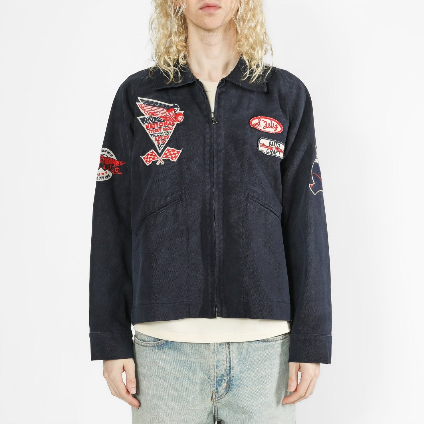 A model with long blonde hair wearing a dark navy blue utility jacket with multiple embroidered patches, paired with light-wash jeans, against a plain white studio background.