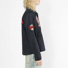 A side-profile view of a model with curly blonde hair wearing a dark navy blue jacket with red and white embroidered patches on the sleeves, paired with light-wash jeans against a plain white background.