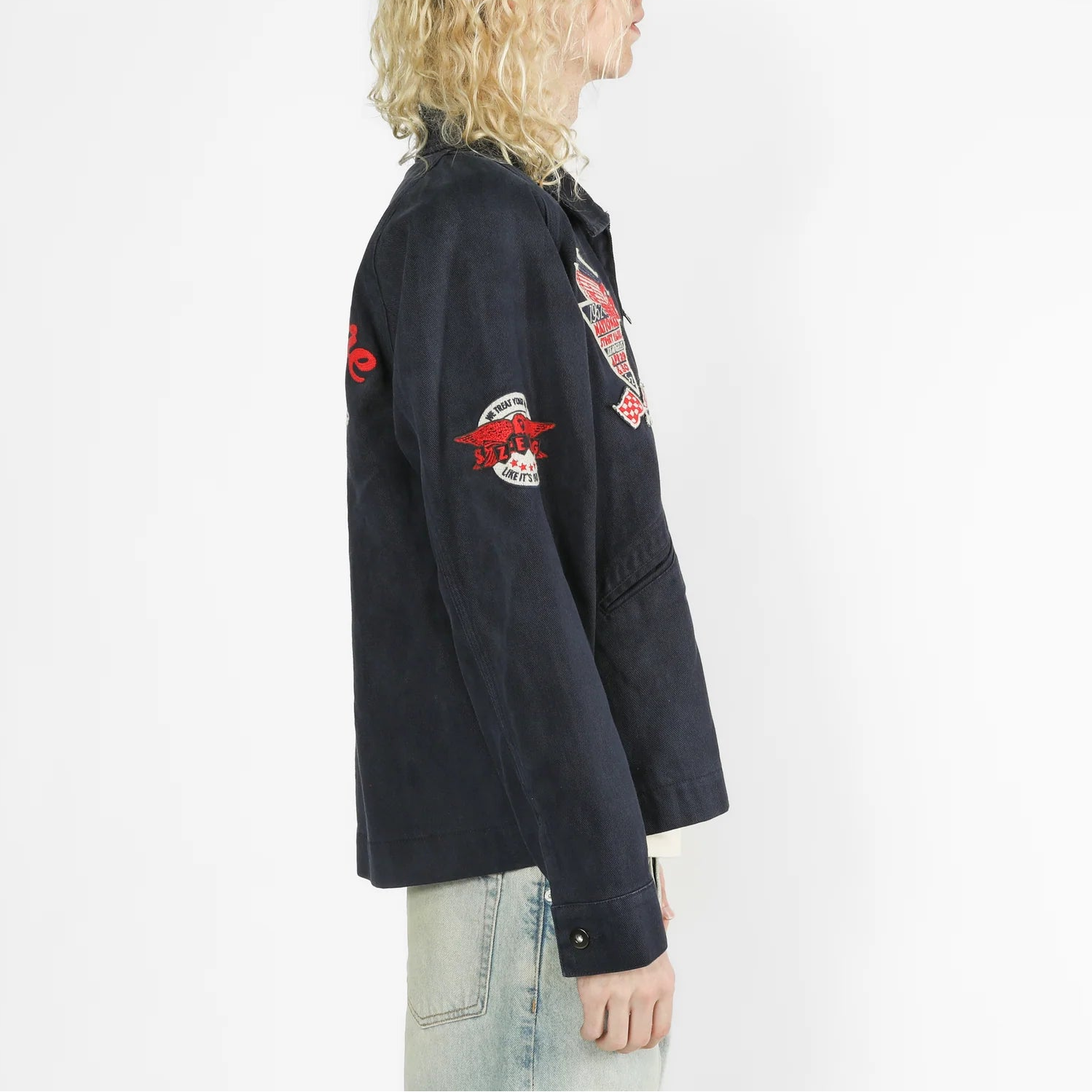 A side-profile view of a model with curly blonde hair wearing a dark navy blue jacket with red and white embroidered patches on the sleeves, paired with light-wash jeans against a plain white background.