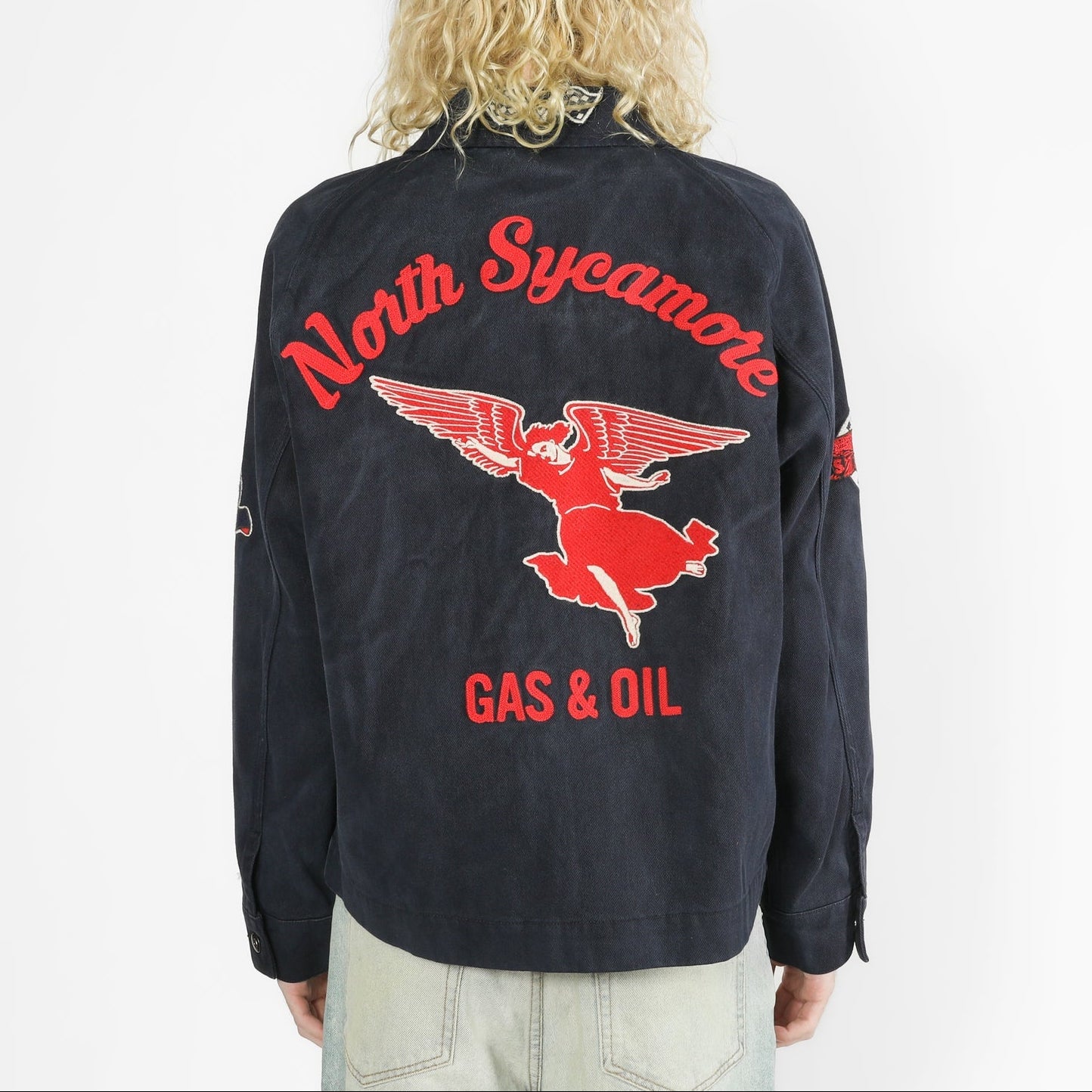 A person viewed from the back wearing a faded black jacket with a large red and white North Sycamote Gas & Oil winged logo graphic on the back, paired with light gray jeans against a white studio background.