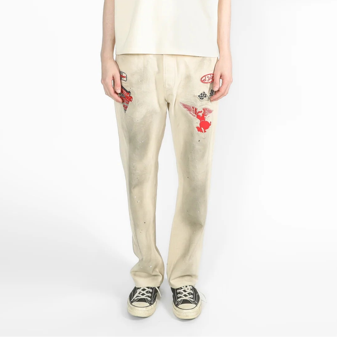A model stands against a white studio background wearing a cream-colored oversized short-sleeve t-shirt, matching cream pants with red embroidered details, and black canvas sneakers.