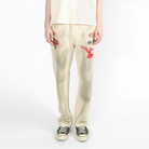A model stands against a white studio background wearing a cream-colored oversized short-sleeve t-shirt, matching cream pants with red embroidered details, and black canvas sneakers.