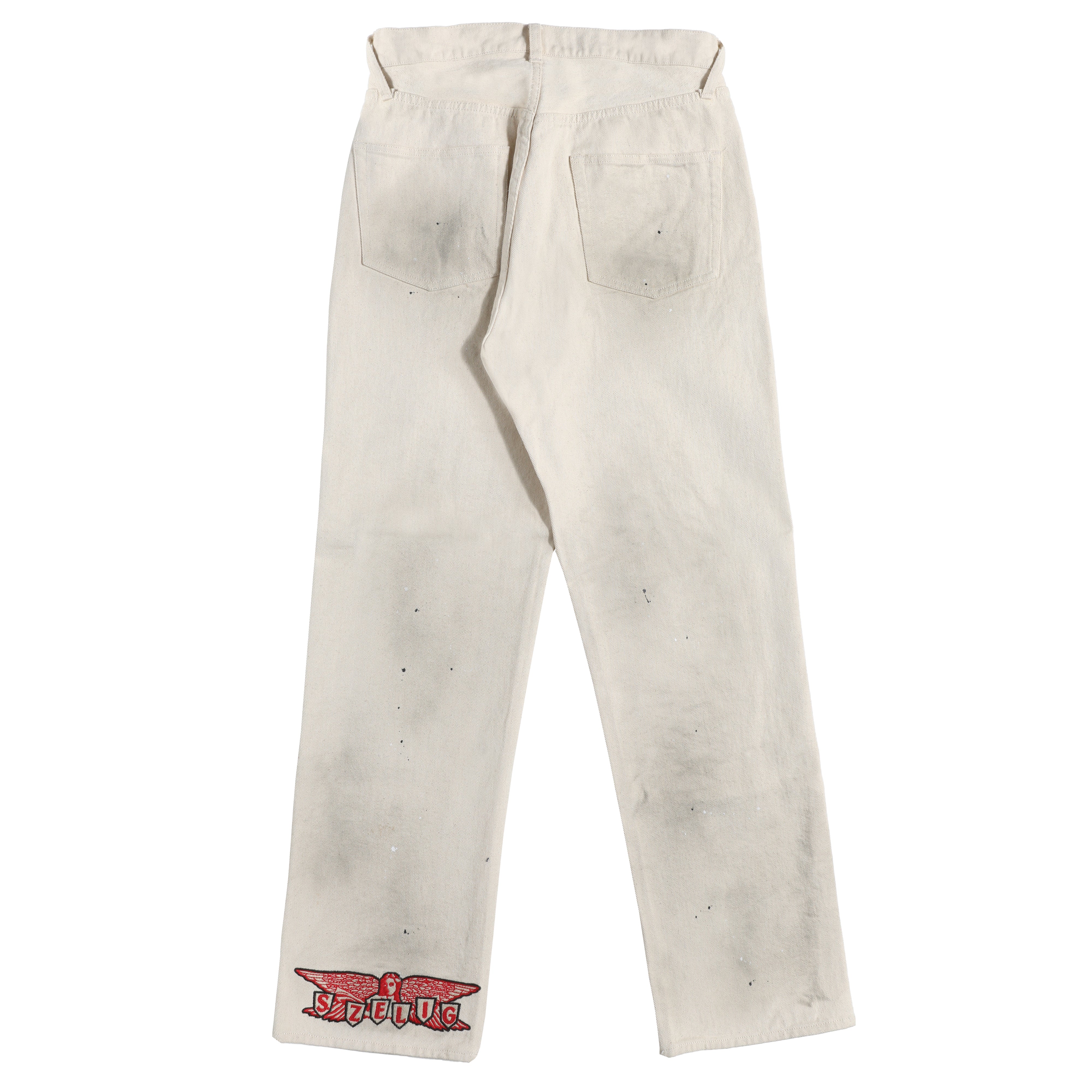 A pair of off-white jeans shown from the back, featuring a red and black 'S ZELIG' graphic patch on the lower left leg and light wash discolorations across the fabric.