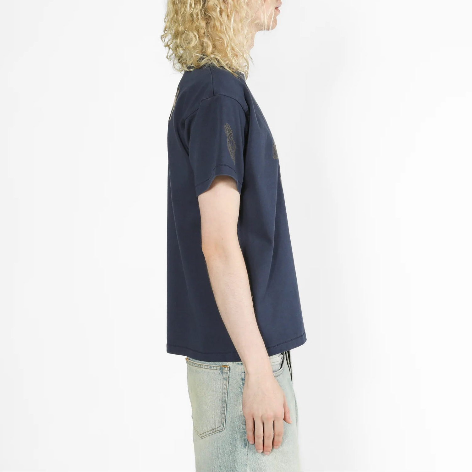 A model with curly blonde hair is shown in profile wearing a dark navy blue t-shirt and light wash jeans against a plain white background.
