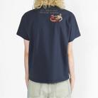 A person viewed from the back wearing a navy blue short-sleeve T-shirt with a small red and beige graphic and text on the upper back, paired with light denim jeans, against a plain white background.