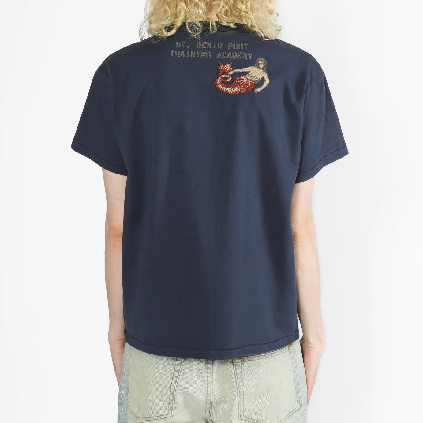 A person viewed from the back wearing a navy blue short-sleeve T-shirt with a small red and beige graphic and text on the upper back, paired with light denim jeans, against a plain white background.