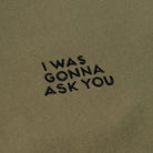 Close-up of a olive green fabric surface with black embroidered text reading 'I WAS GONNA ASK YOU'.