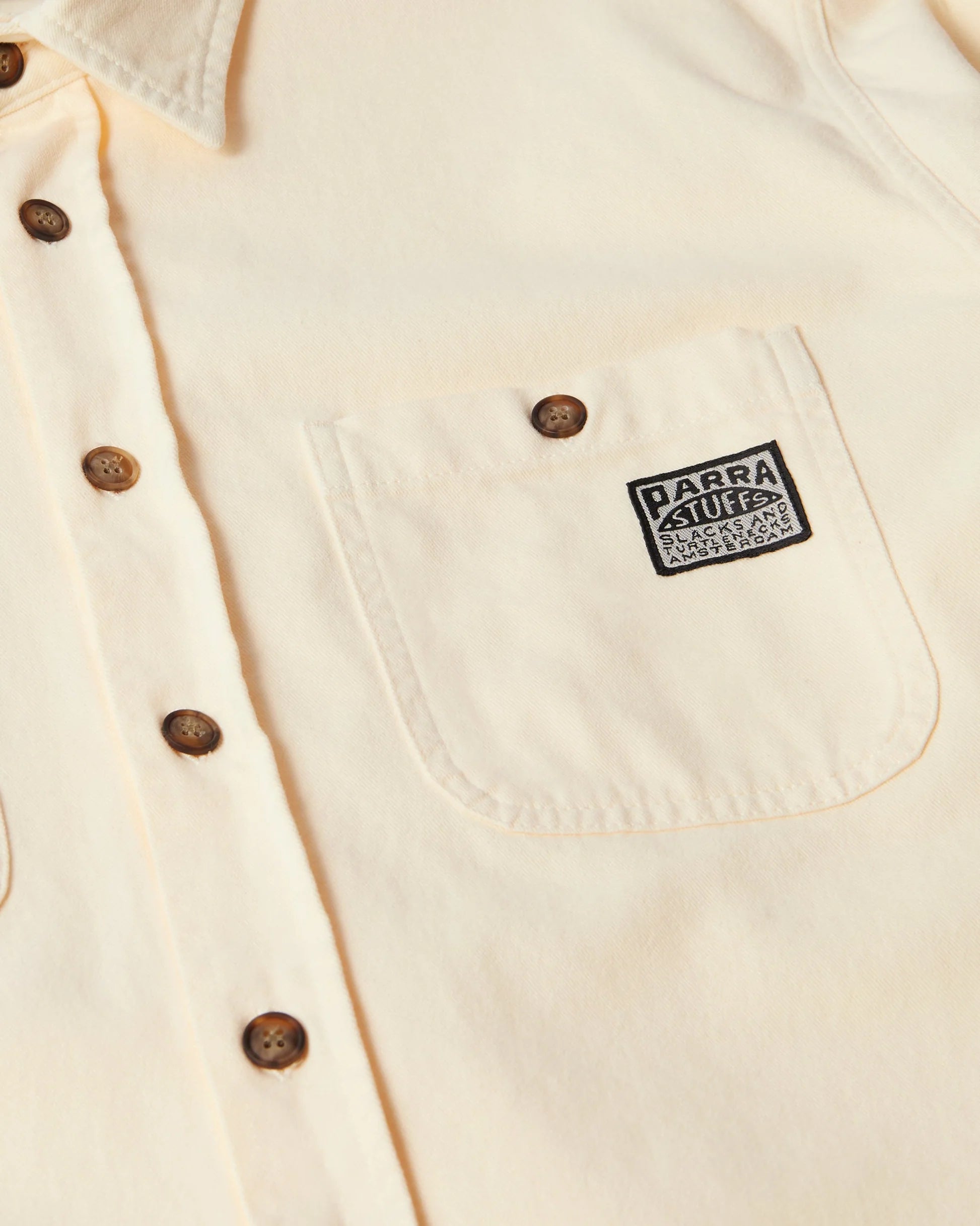 Close-up of a cream-colored button-down shirt with a chest pocket featuring a black and white 'Parra' patch.