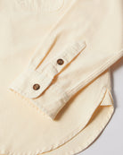 Close-up of a cream-colored long-sleeve shirt showing the buttoned cuff and part of the curved hem, with two brown buttons on the cuff.