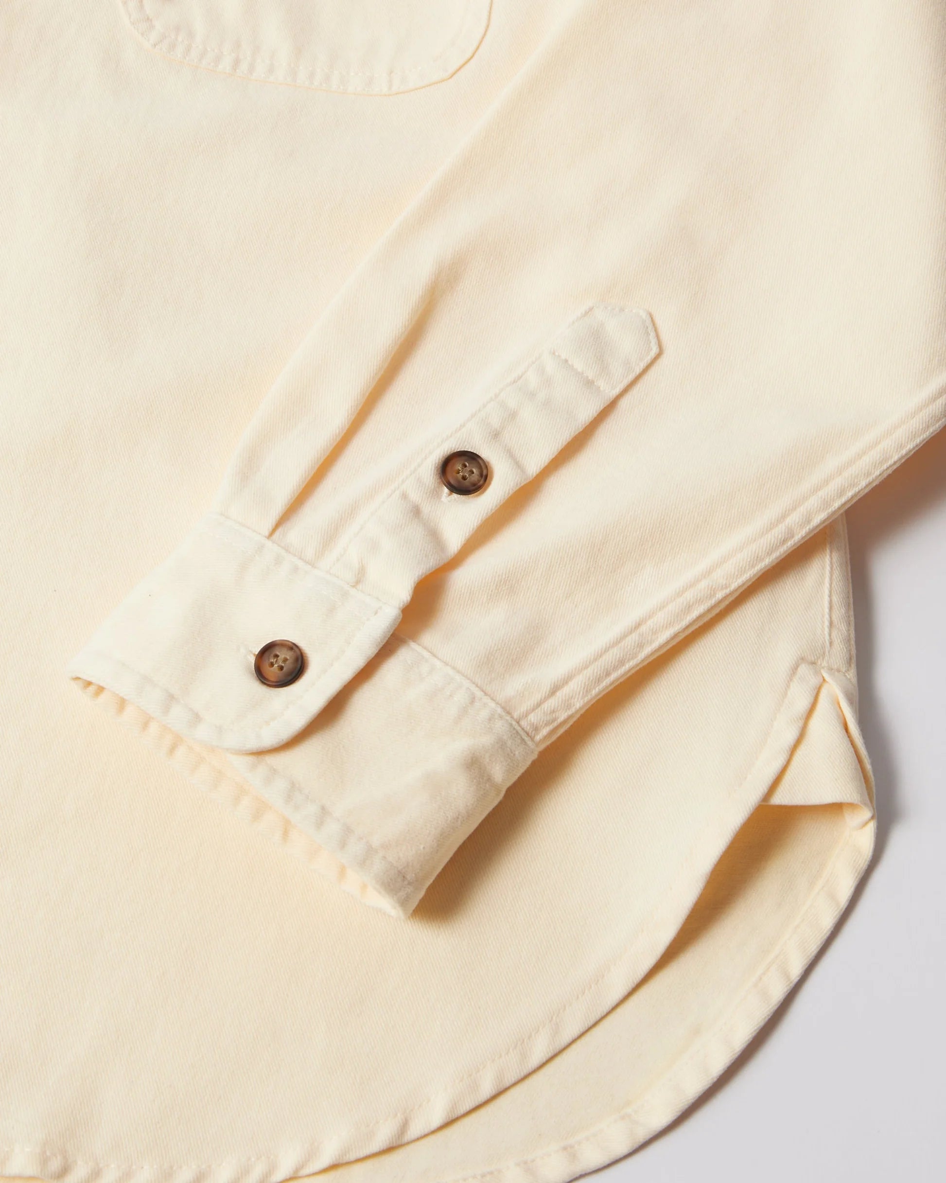 Close-up of a cream-colored long-sleeve shirt showing the buttoned cuff and part of the curved hem, with two brown buttons on the cuff.